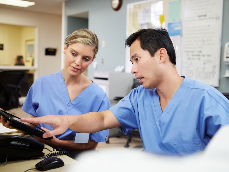 Two healthcare professionals in blue scrubs engaged in a discussion over a digital tablet in a clinical setting.