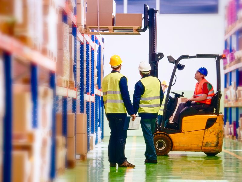 Two workers in high visibility vests and hard hats are observing while another operates a forklift in a warehouse.