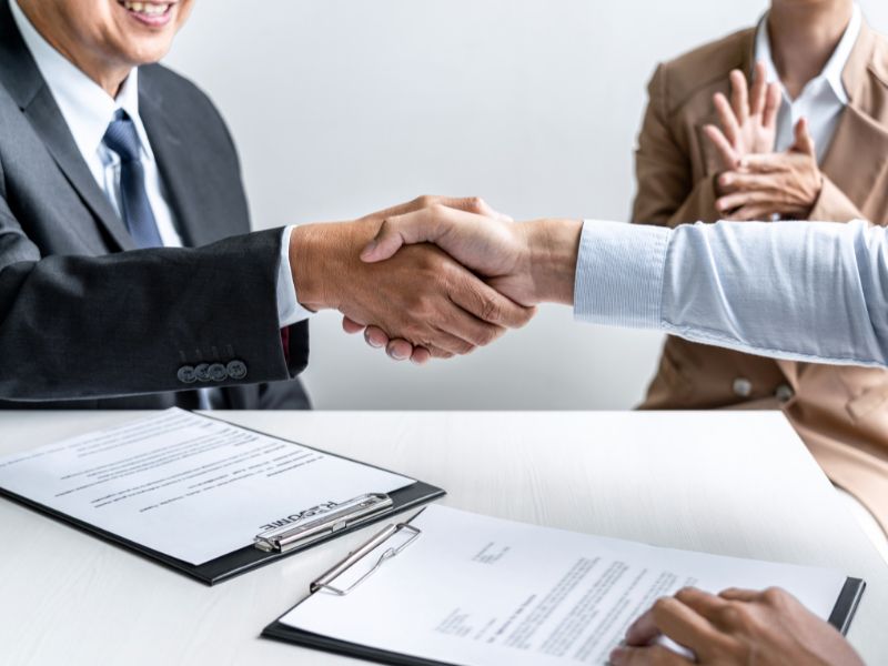A handshake between two individuals in professional attire, with a contract on the table and another person in the background, indicating a business agreement.