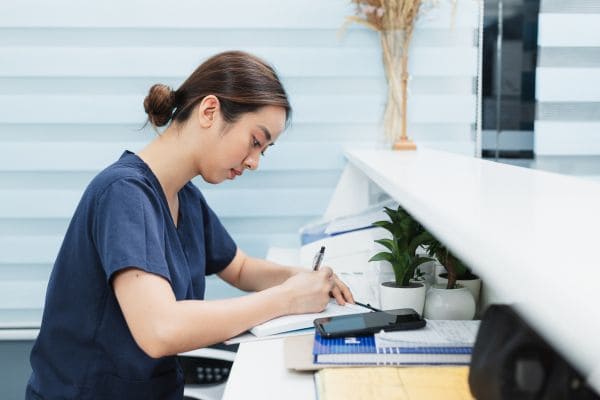 Nurse in scrubs writing on a clipboard at a clinic reception desk.