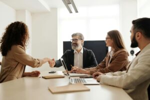Group of business people in a meeting room, two shaking hands while others observe.